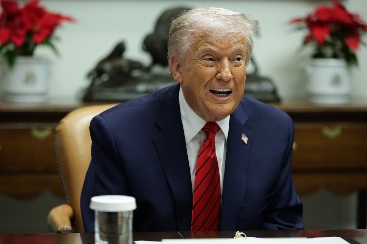 An elderly man in a suit speaks as he sits. He has an American flag pin on his lapel.