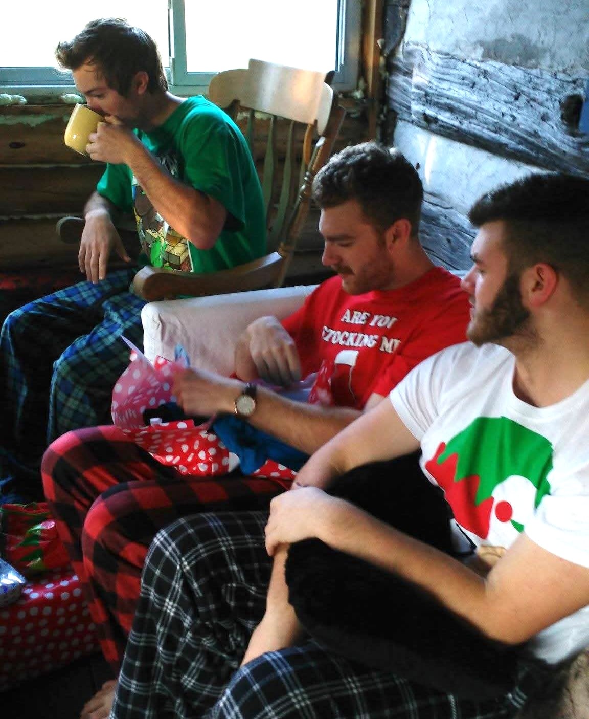Three young men wearing holiday shirts and pyjama pants open presents.