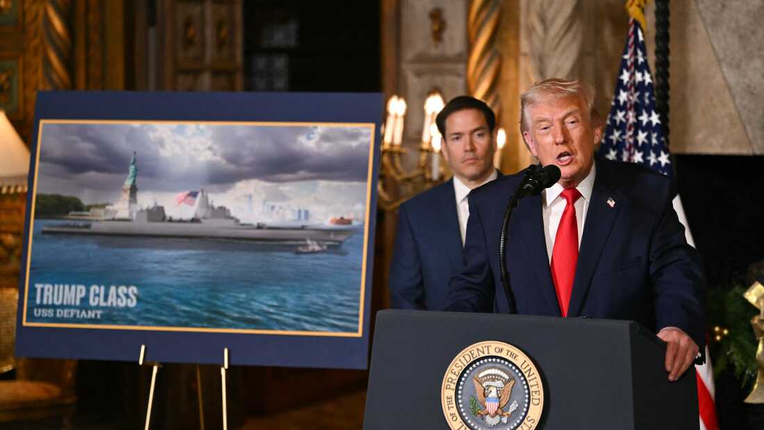 President Trump talks at a podium as he announces a new class of warship. U.S. Secretary of State Marco Rubio stands behind him on his left side. To the left of both of them is a mock image of the class of warship seen in front of the Statue of Liberty.