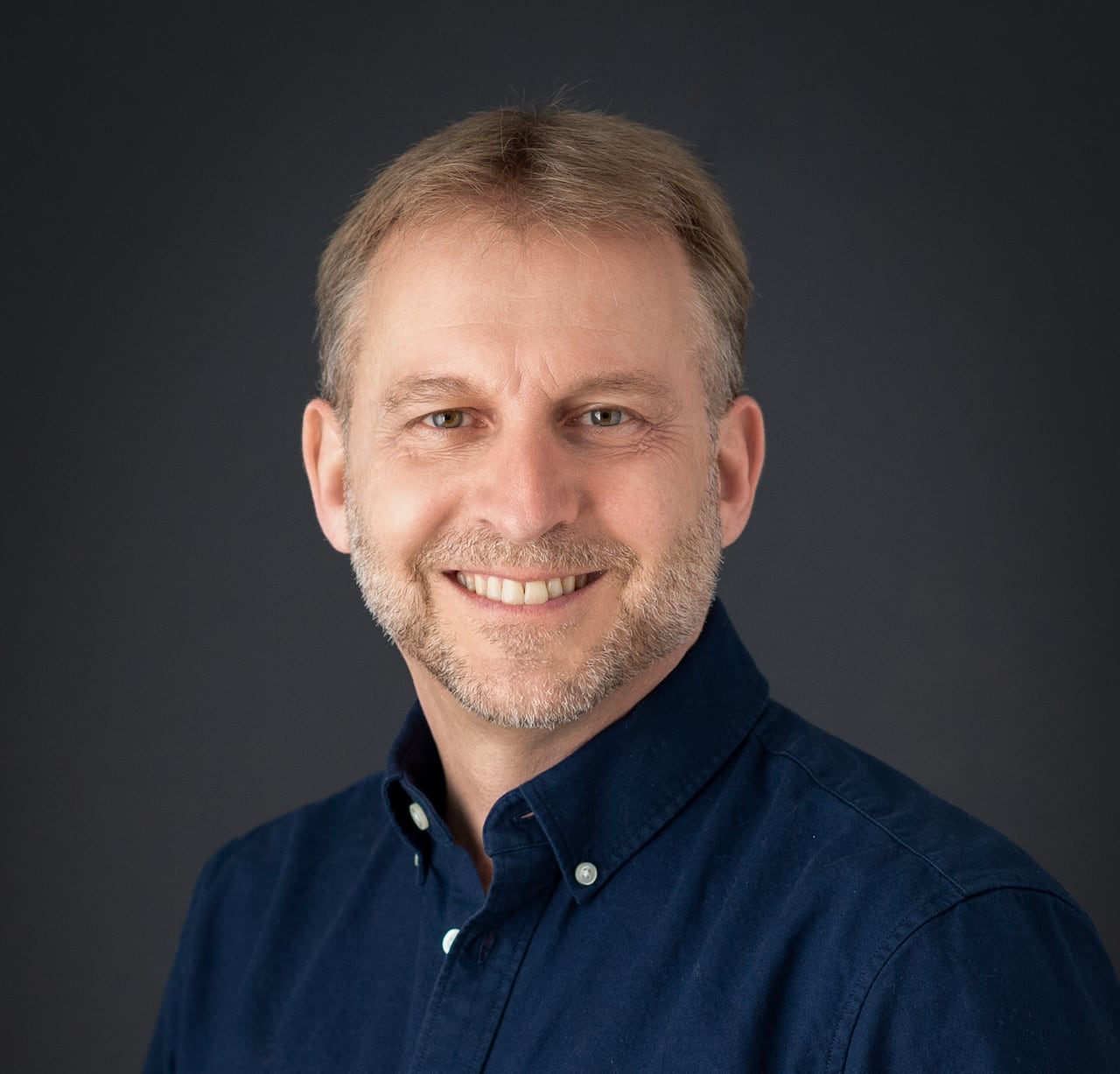 Man in dark blue collared shirt smiling in front of dark backdrop