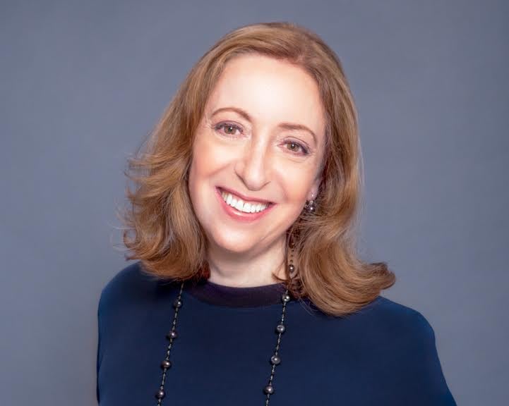 Woman wearing navy short-sleeved top and a necklace stands in front of dark background