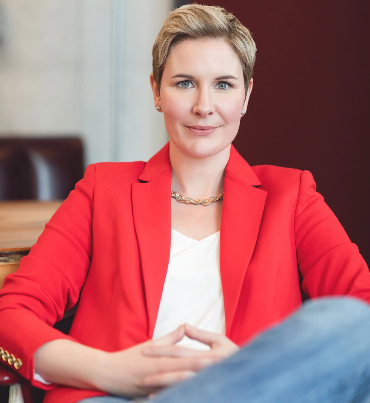 Woman with short hair, red blazer and jeans sits cross-legged