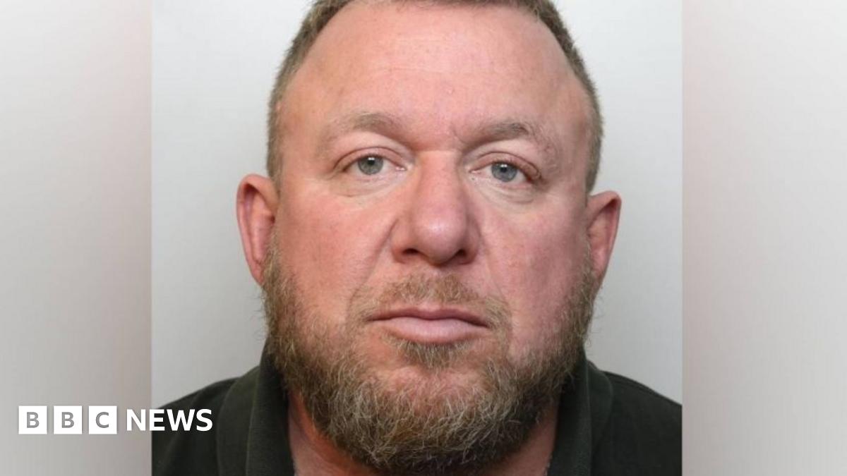 A police custody head and shoulders mugshot of Mark Killick. He has a scruffy dark brown beard and receding hair on his head and is wearing a dark coloured polo top
