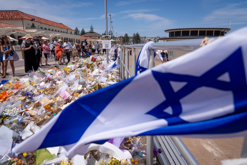 A large number of bunches of flowers are laid across a walkway at Bondi Beach. A Jewish flag flies alongside them.