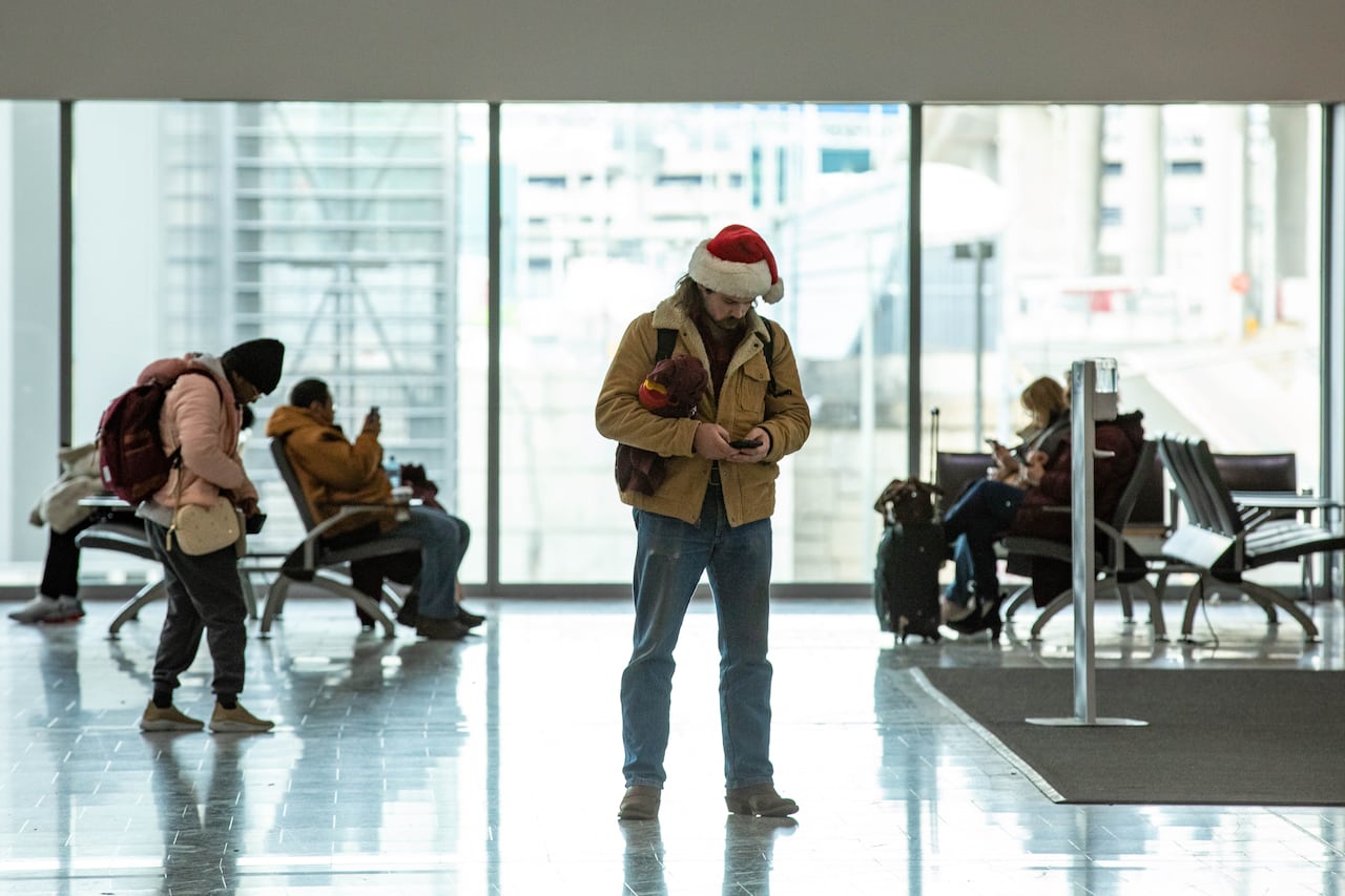 A man in a brown jacket and blue jeans, with a red and white Santa hat on his head, looks down at a phone in his hand while standing in the passenger area of an airport.
