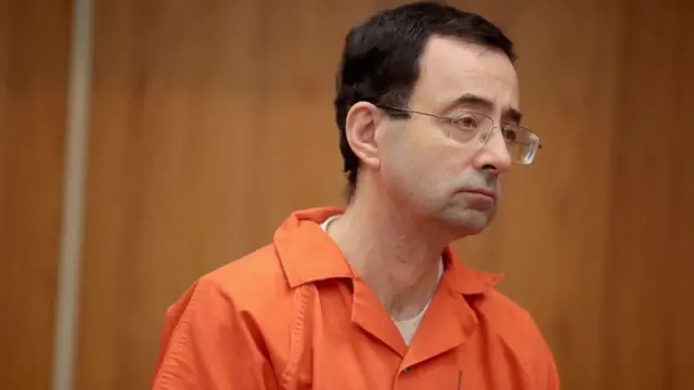 Larry Nassar from di shoulders up, wear square framed glasses wit wire rims and orange jumpsuit, e dey look to im left