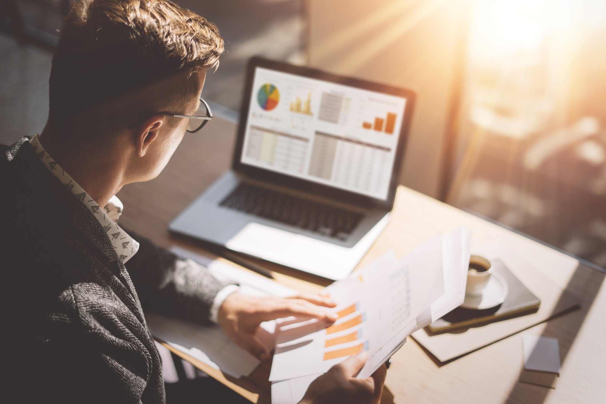 A person holding paper documents looks at a computer as sunlight spills through window.