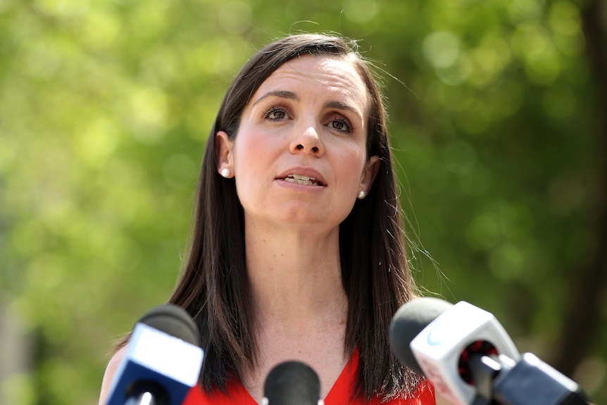 Courtney Houssos speaks in front of three microphones. Behind her are trees. She is wearing a red shirt.