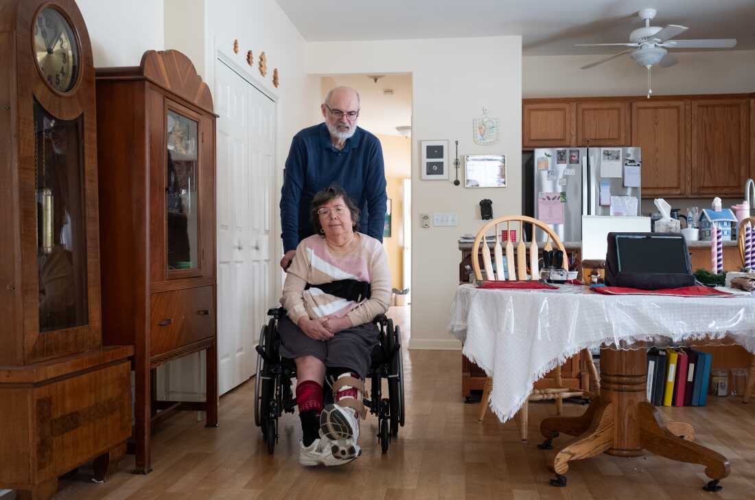 A man with a gray beard has his arms on the back of the wheelchair of a woman with short hair. They are in a dining room with elegant wood furniture and a table covered in a white table cloth.