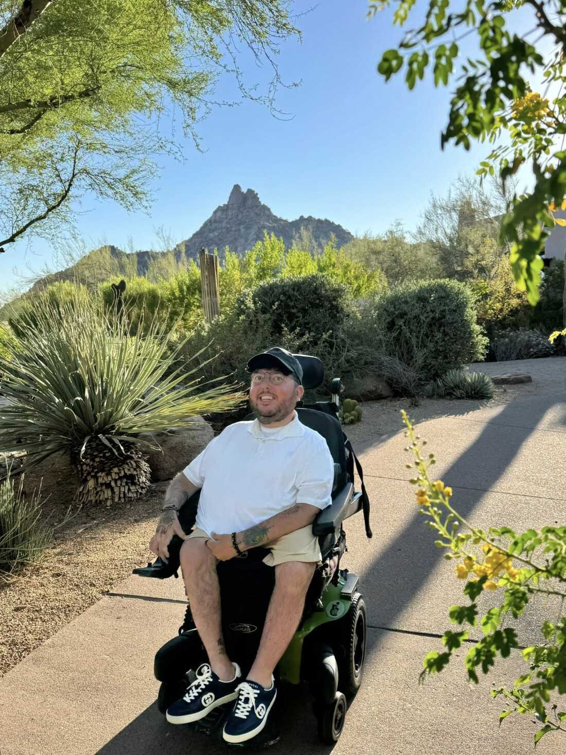 A young man in a white shirt and shorts is outside flanked by trees and greenery. A mountain looms in the distance.