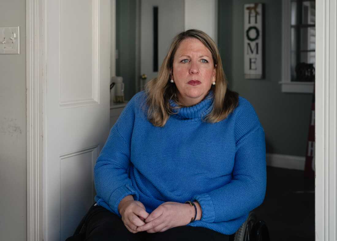 A woman with blonde hair and a blue sweater sits in a wheelchair. On the back wall is a vertical sign that says: HOME.