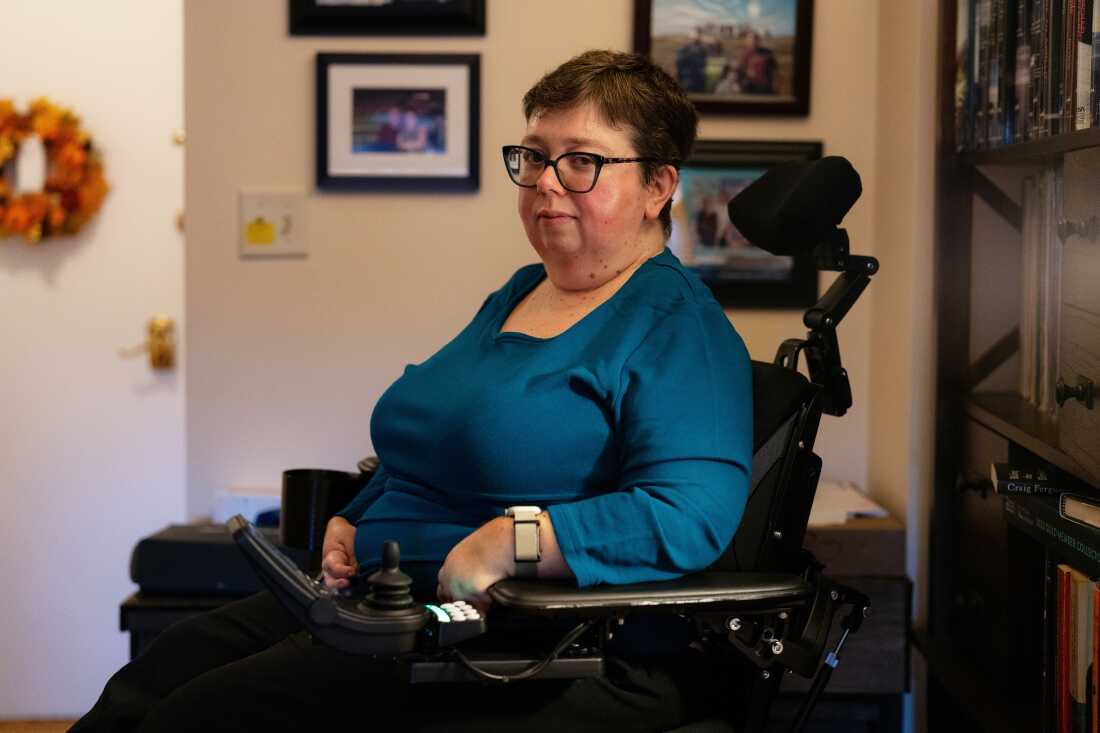A woman with short hair wearing a blue top sits in a wheelchair at home. On the walls are photos.