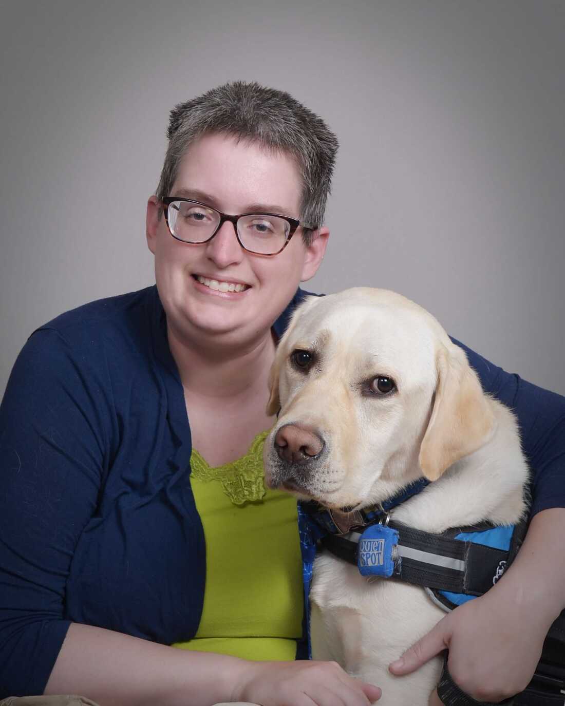 A woman with short hair and glasses smiles as she hugs a service dog, a yellow Labrador Retriever.