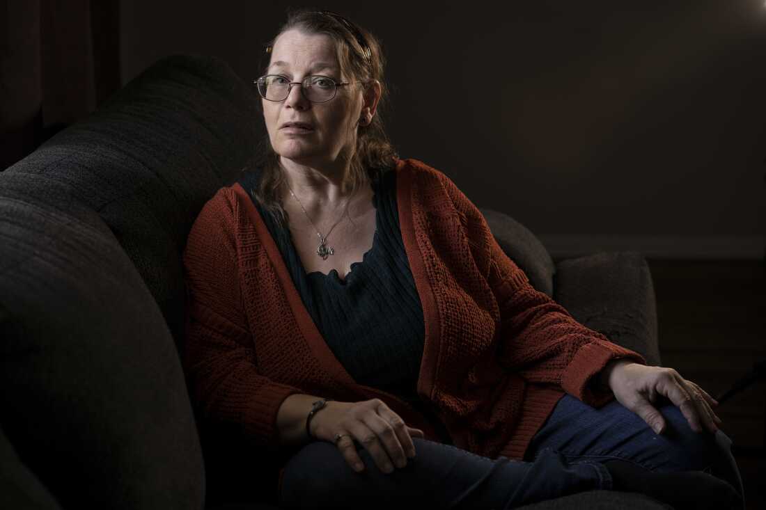 In front of a dark background, a woman with short hair and glasses in a reddish sweater and dark top, in a wheelchair, looks pensively at the camera.