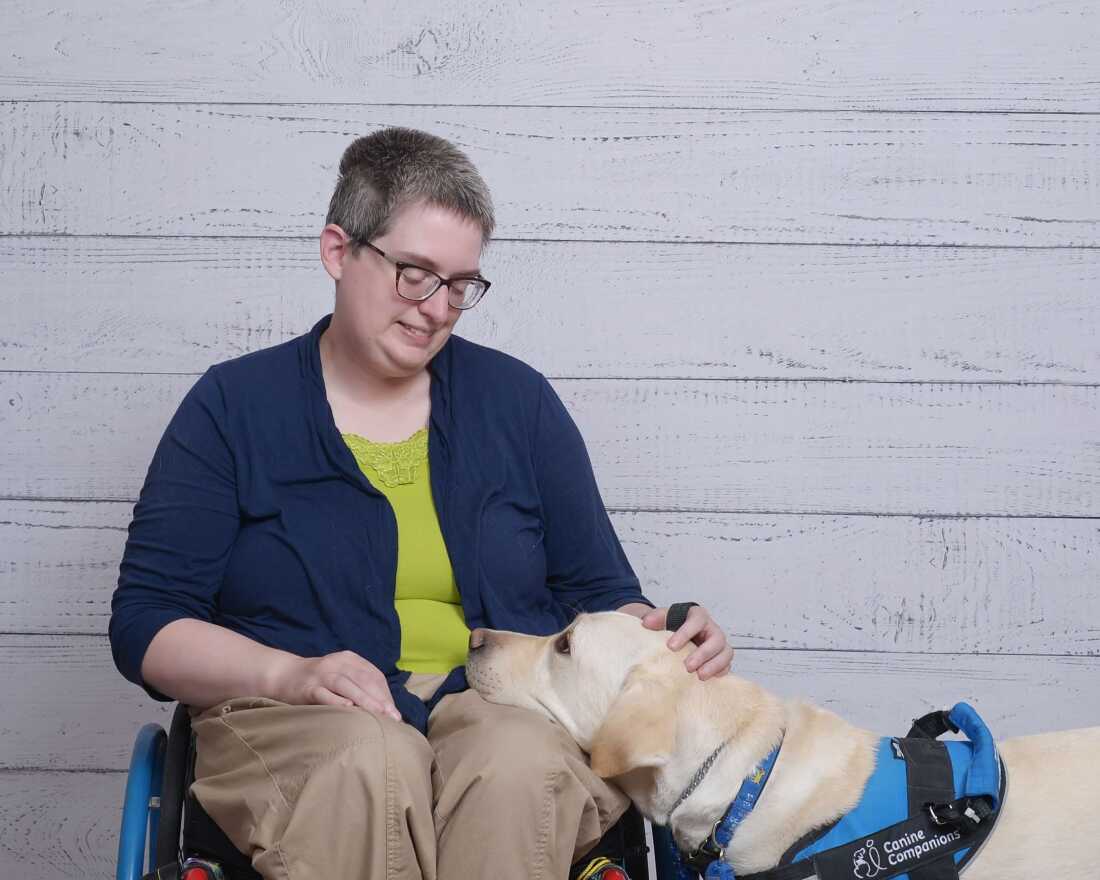 A woman with short hair and glasses, in a wheelchair, pets the dog, a Labrador Retriever, that puts its head in her lap and looks up at her.
