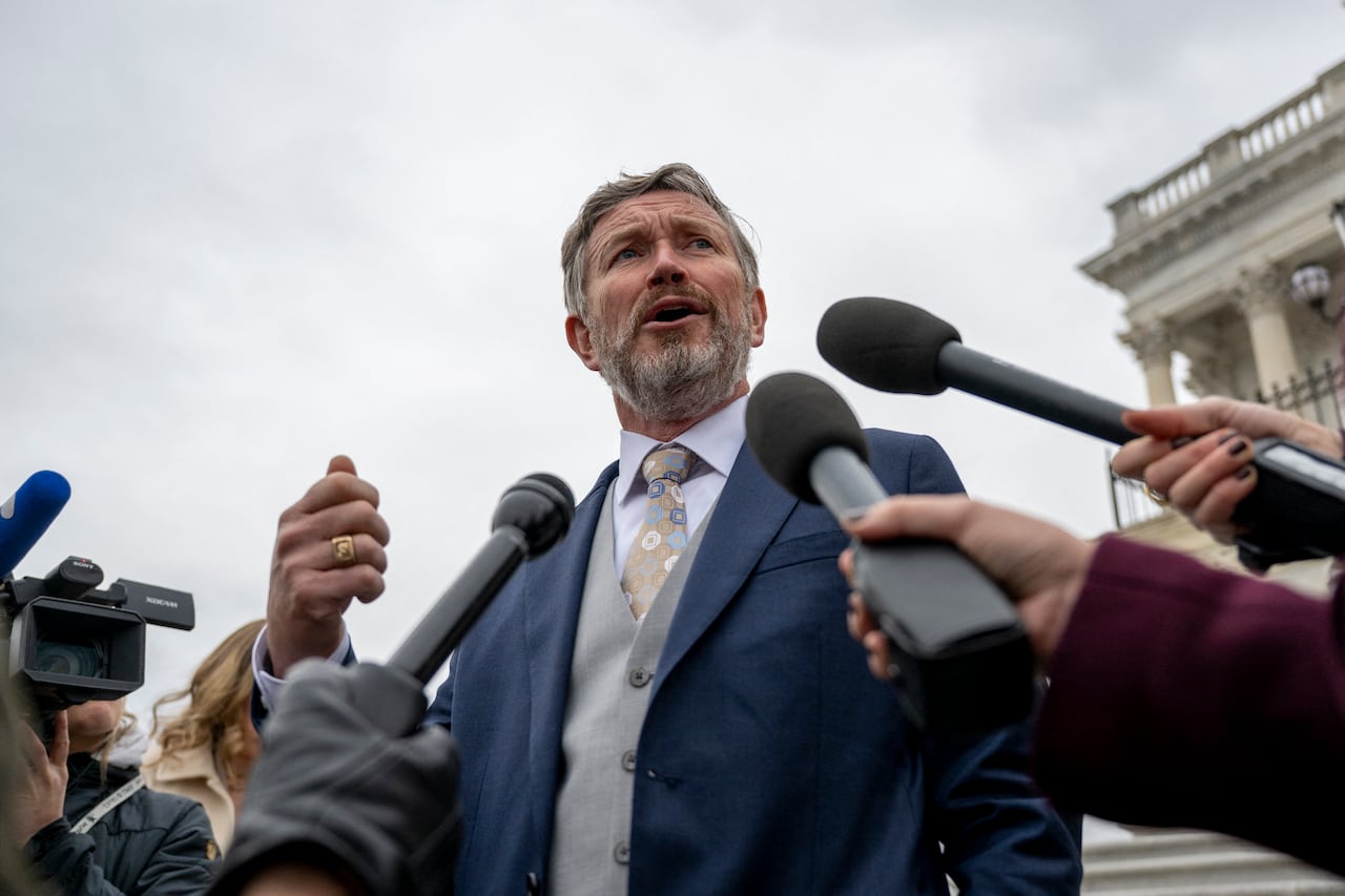 A man in a blue suit speaks to reporters holding microphones at him.