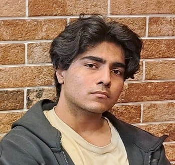 Brunette man with beige shirt and grey zip-up in front of brick wall