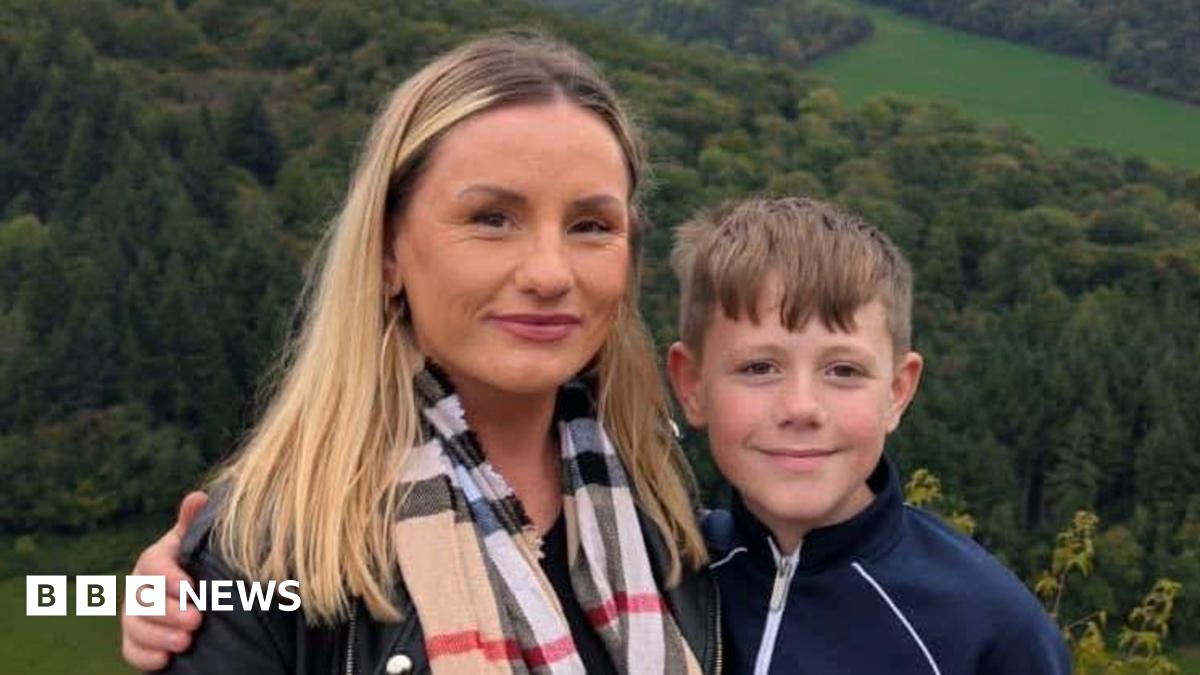 Nicola and Zac standing side by side. They are standing in front of a scenic view with a forest and field behind them. They are both smiling at the camera and Zac has his around around Nicola.