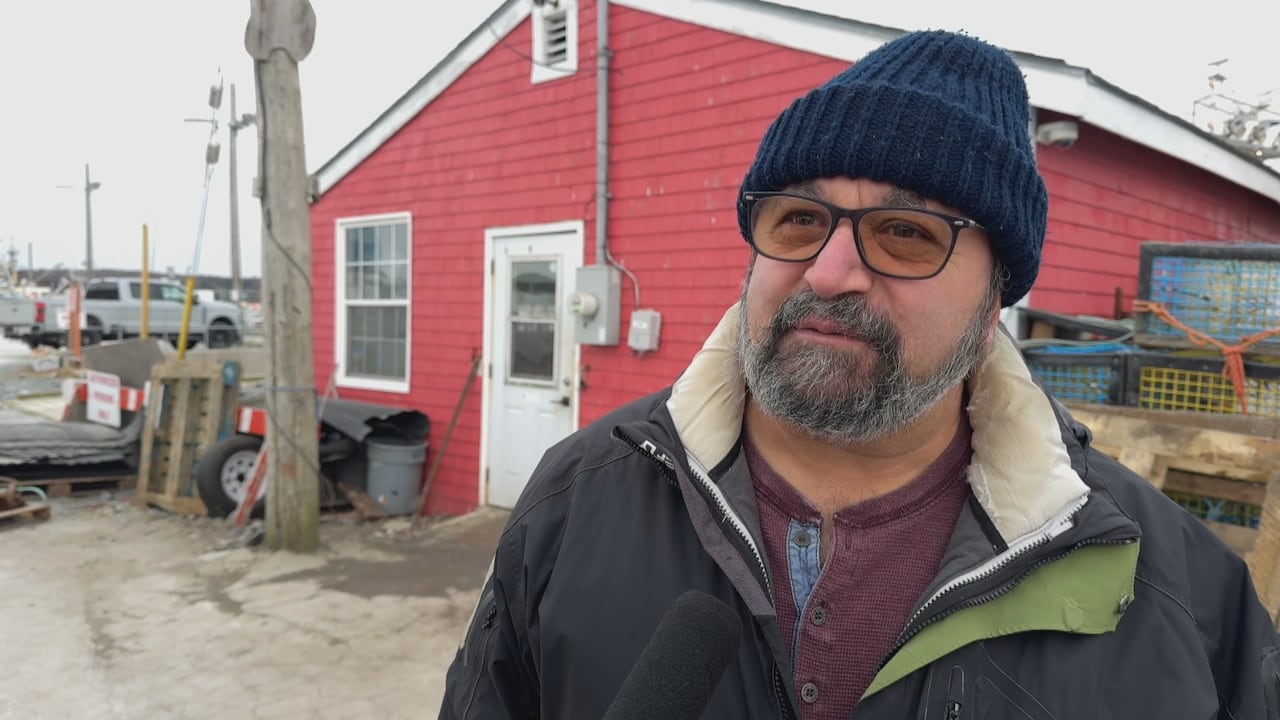 A man wearing a toque and wearing glasses is shown outside in front of a red building.