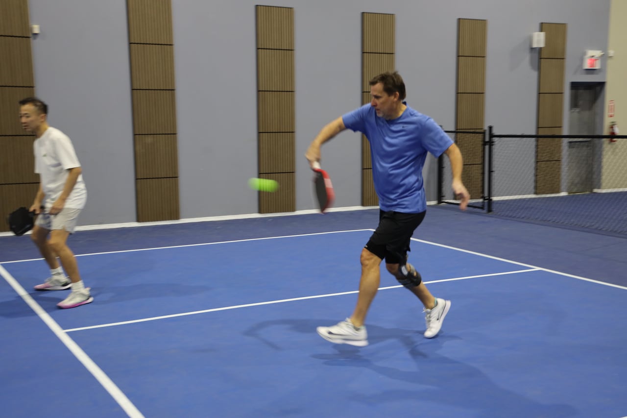 A man in a blue t-shirt hits a pickleball with a paddle.