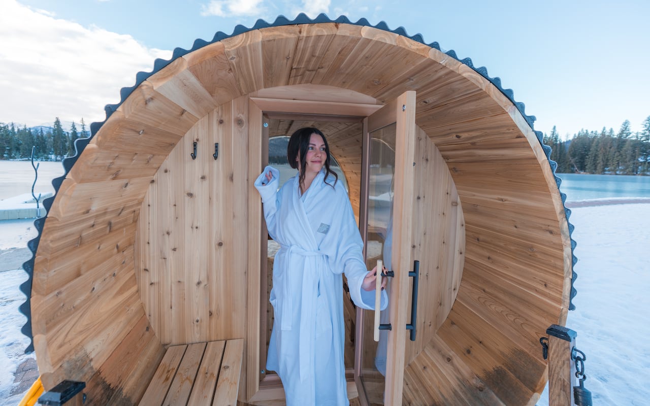 A woman in a white robe emerges from a barrel sauna into a winter landscape of a frozen lake and snow