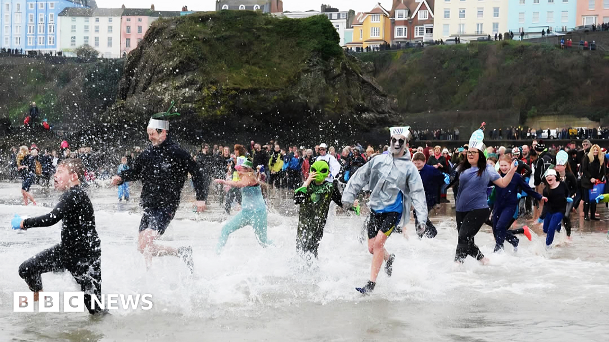 A large crowd of people, some in fancy or Christmas dress, is running into the sea. There is a large moss-covered rock on the beach behind them and a raised walkway around the beach with people standing spectating. A grassy cliff frames the beach with a street above it, backed by blocks of brightly painted townhouses in different colours.