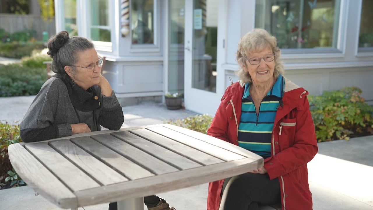 Two elderly women sit together at an outdoor table smiling.
