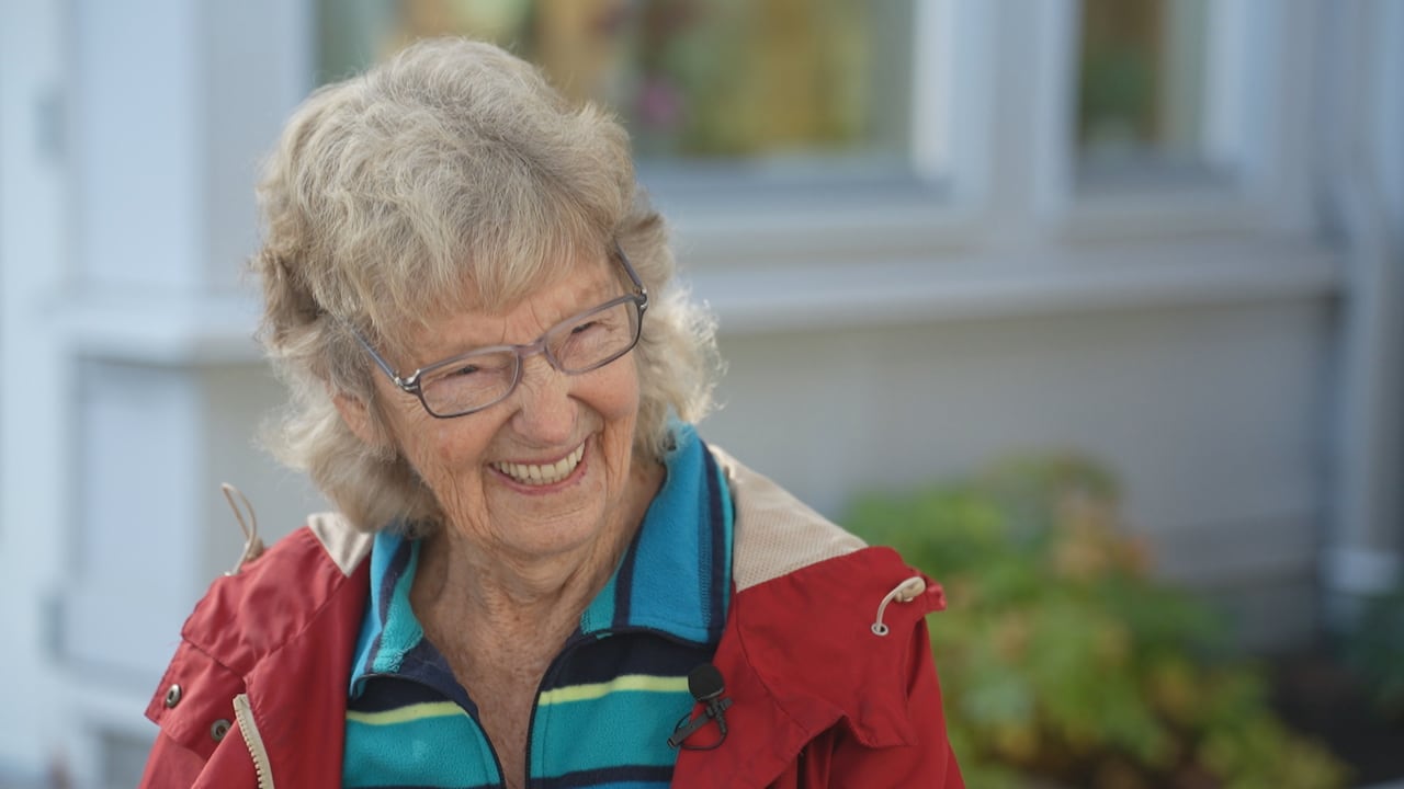 A close-up of an elderly woman smiling outdoors, wearing glasses and a red jacket.