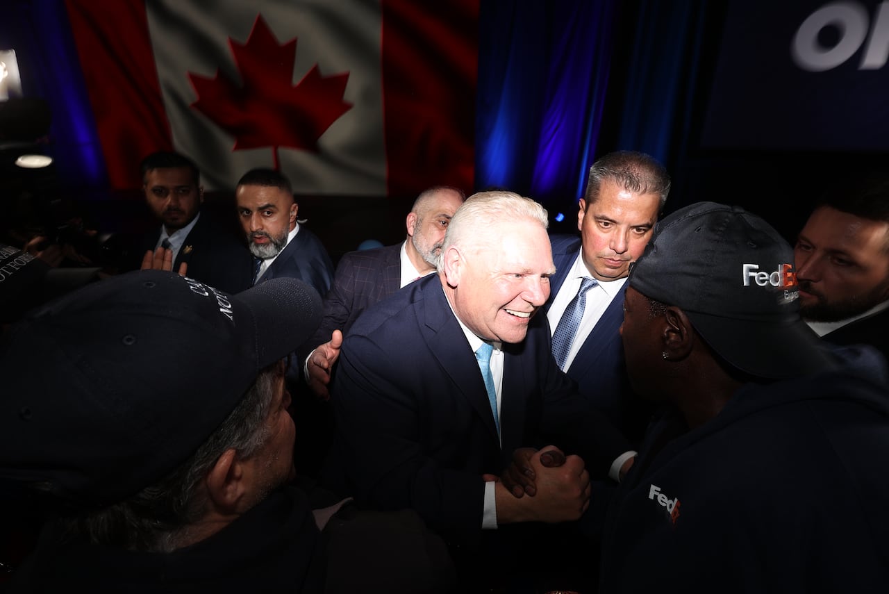 White-haired politician in suit and tie shakes hands with a supporter in baseball cap inside a congress centre.