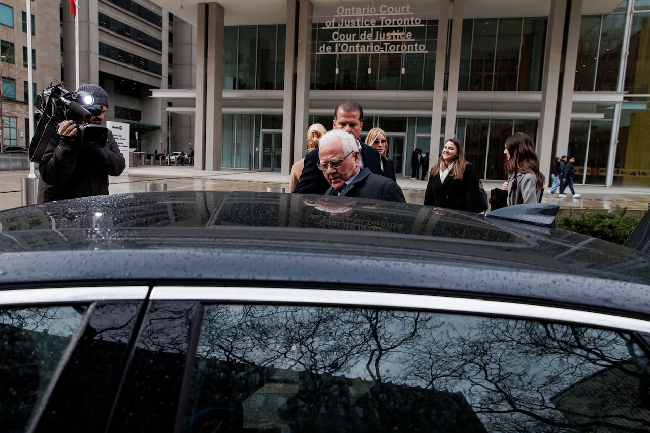 An elderly man about to get into a chauffeured vehicle, with a camera operator at the front of the vehicle filming while a group of people stand behind him.