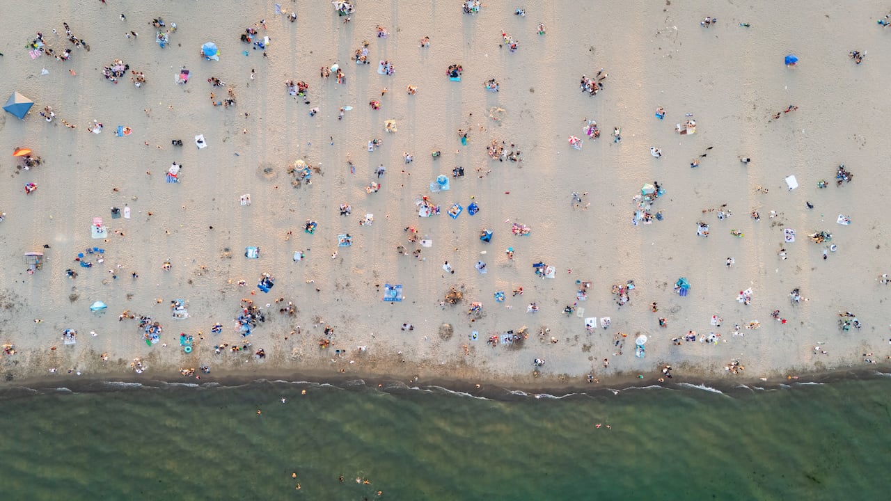 A bird's-eye view of sunbathers on a beach near a shoreline shortly before sunset, with soft warm gold light.