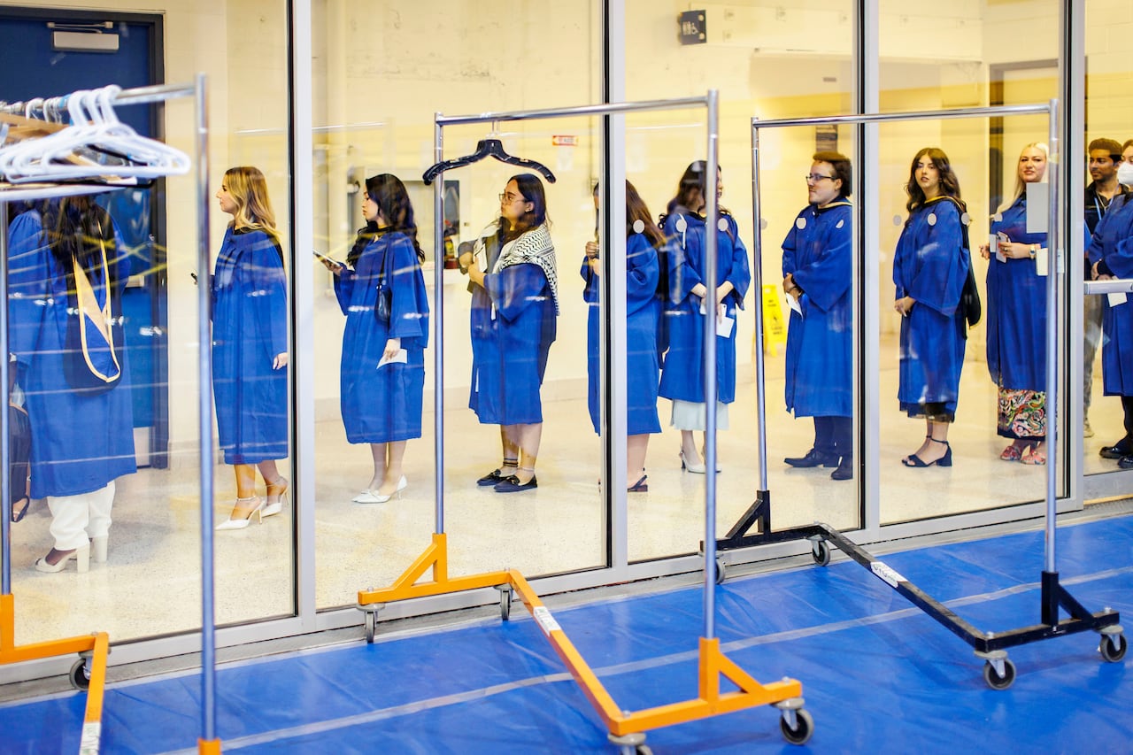 Students in their regalia in a line seen near floor-to-ceiling windows with a view of coat racks opposite them.