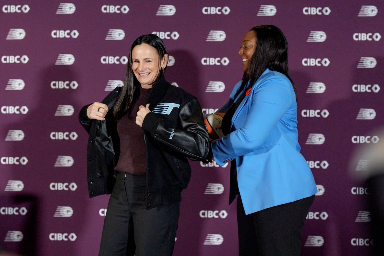 A woman putting a bomber jacket on another woman, standing in front of  wallpaper that reads: CIBC.