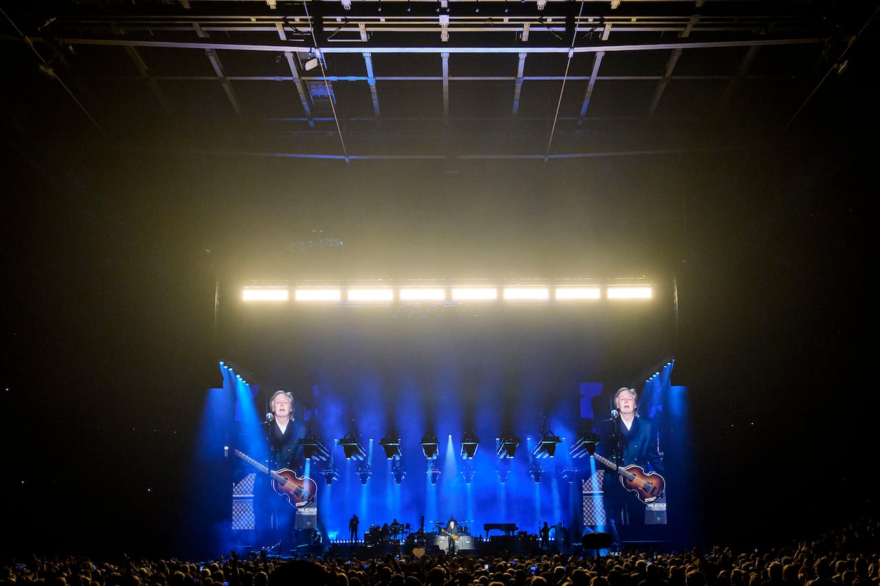 A singer on stage lit in blue stage lights, along with other musicians, and packed audience in the foreground, seen from back of the concert stadium.