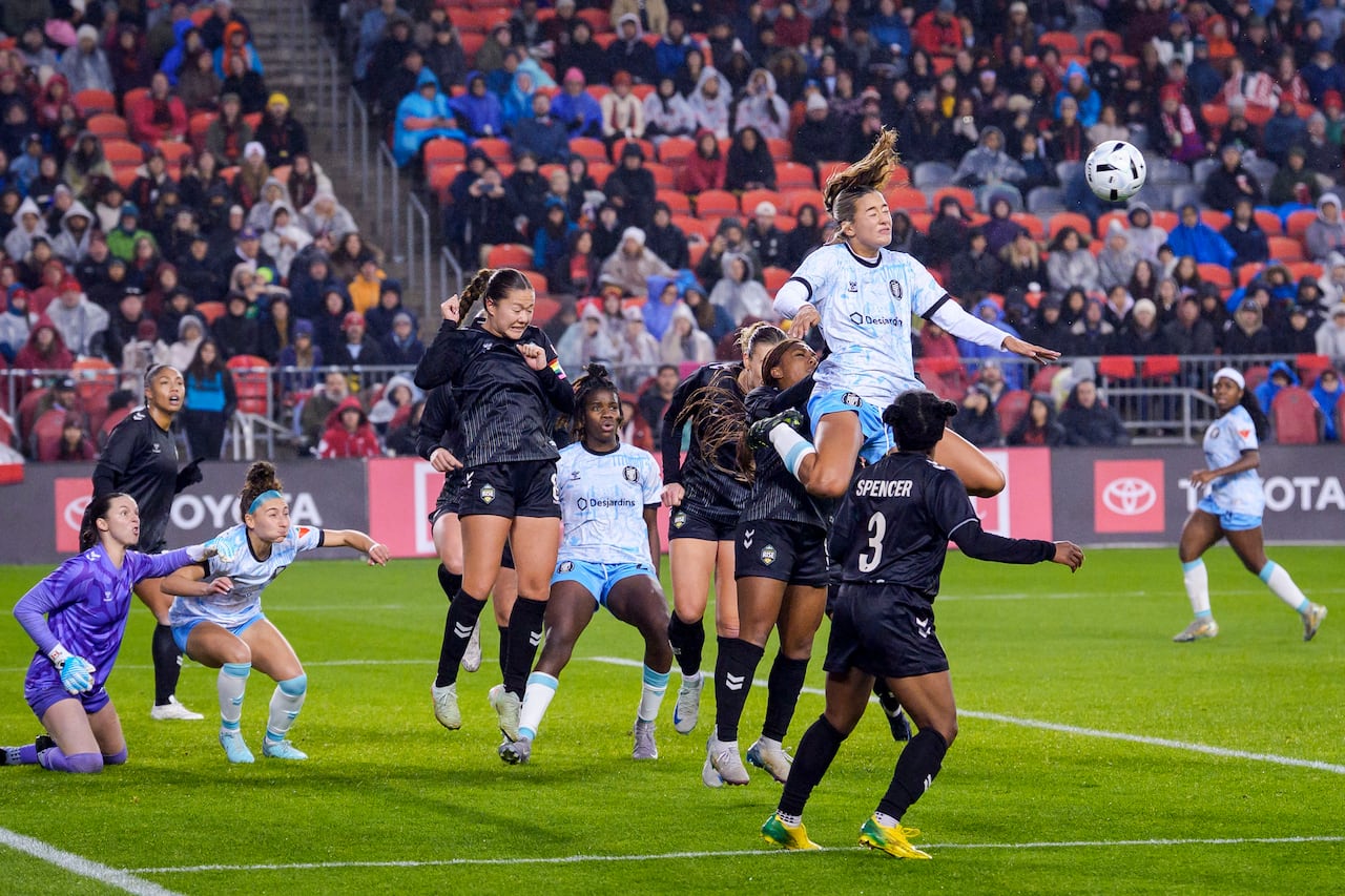 A soccer player in mid-air doing a header during game action against rival players on the field, with spectators in the bleachers in the distance. 