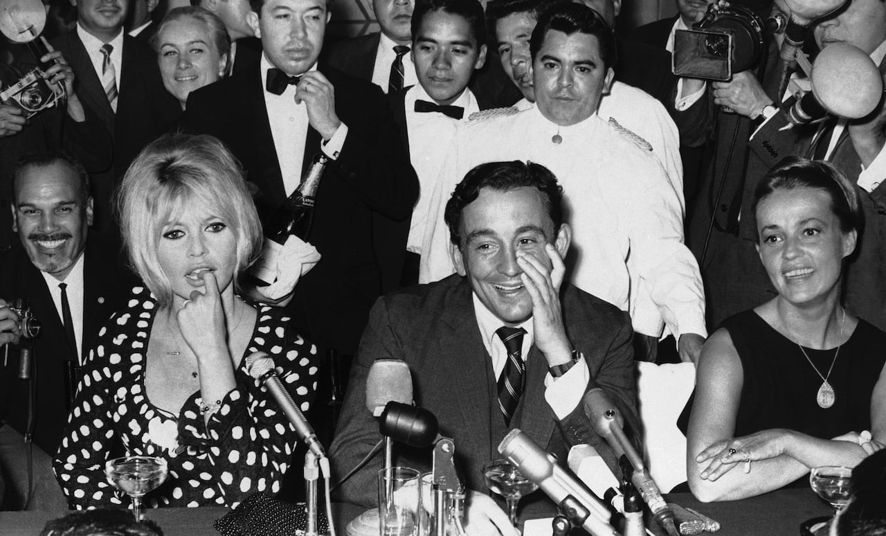 Two women are seated on either side of a man at a table with microphones in a black and white photograph.