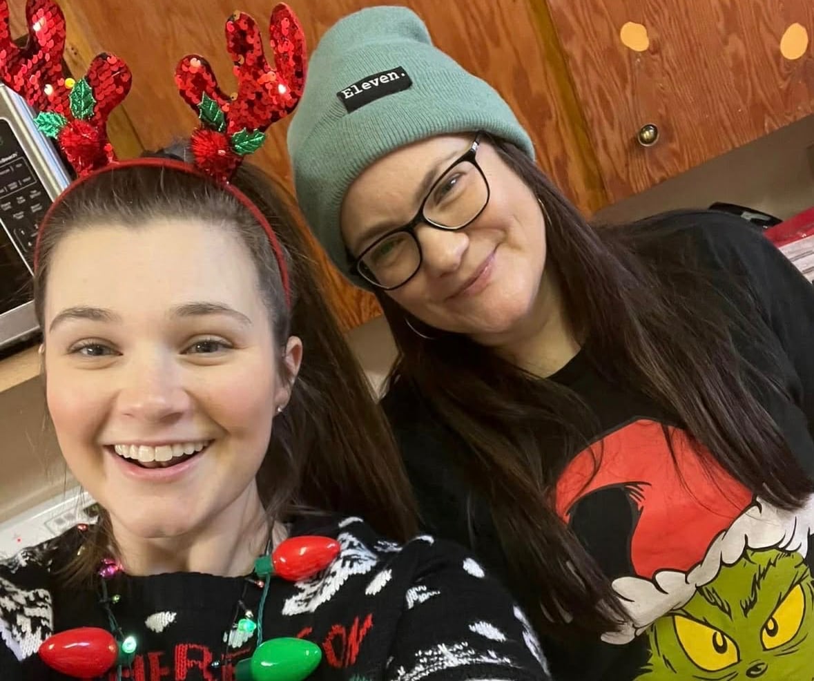 A white woman wearing a Christmas sweater and antler hairband, she is standing next to her mother who is wearing a Grinch sweater, glasses and a hat.
