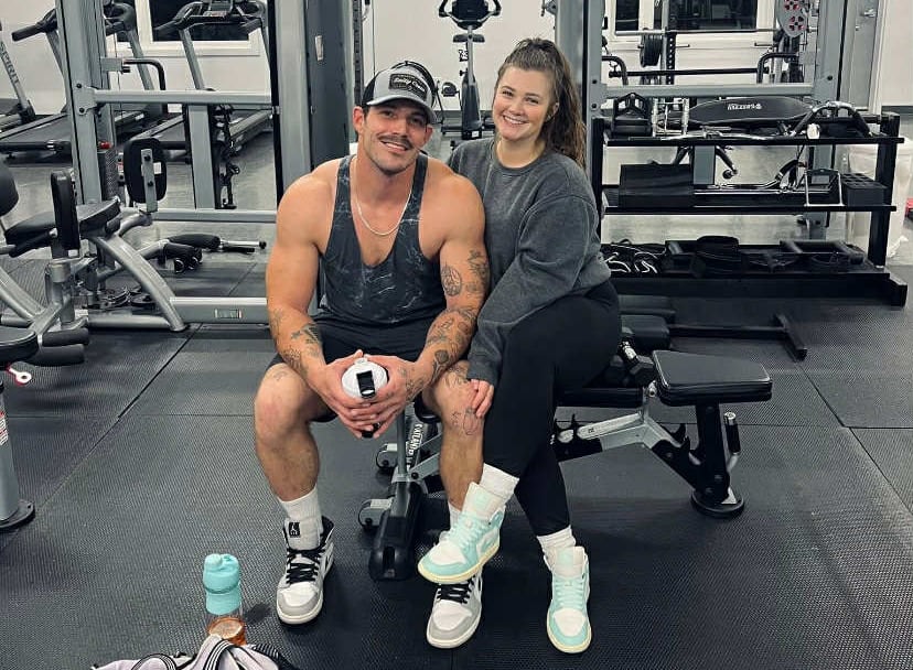 A white muscular man with a white woman wearing a hoodie, they are both sitting on a bench, close to each other, in the gym. 