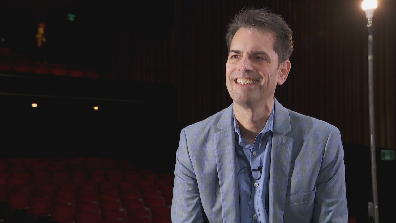 A man smiles wide in front of the backdrop of a theatre with red velvet seats. 