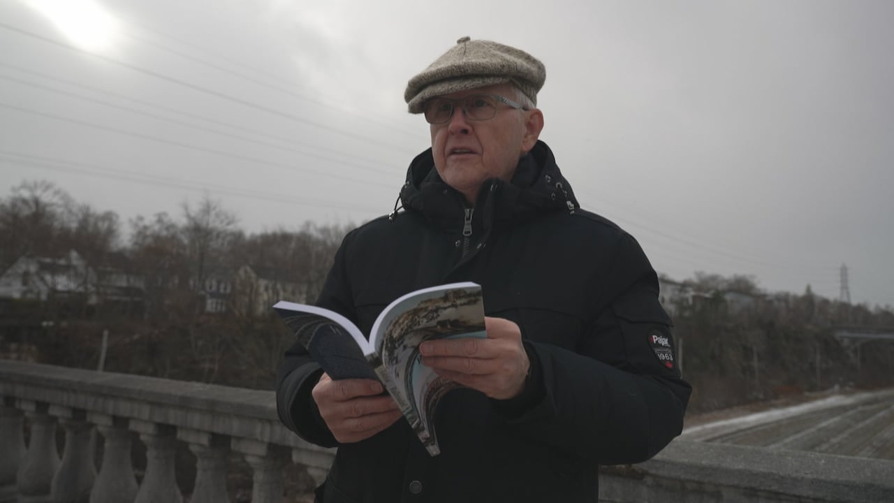 A man in a tam o' shanter, holds a book while standing on a bridge over the rail cut