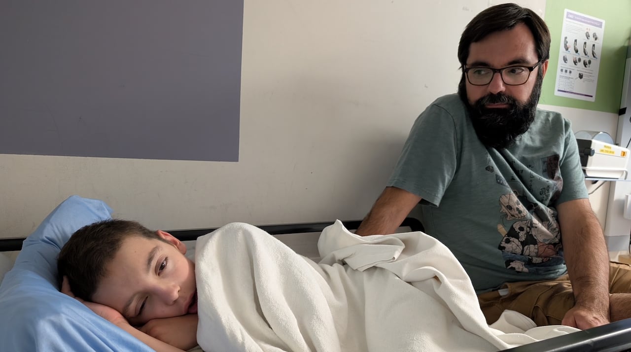 A boy lies in a hospital bed, his dad sits on the bed next to him.