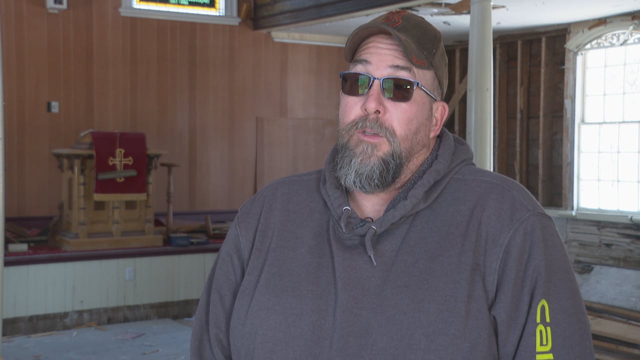 A white man with black and white French beard. He is wearing a hat and brown sunglasses.