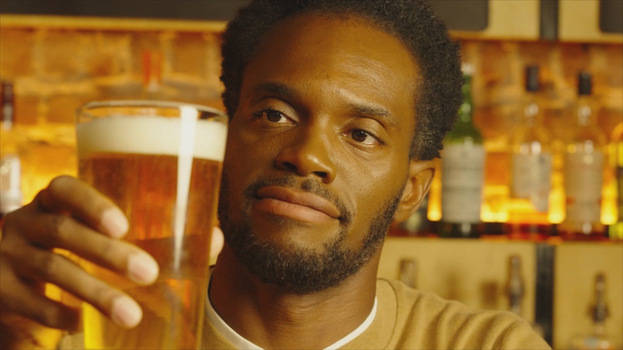 A close-up, warm-toned shot of a man with a short beard looking thoughtfully at a tall glass of beer he is holding up.