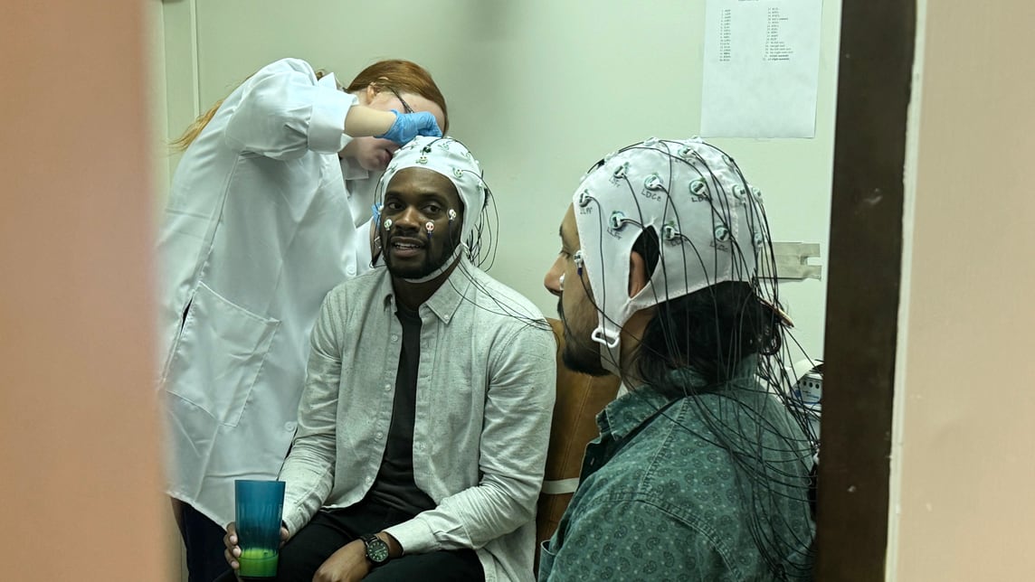 A medium shot captures two men seated in a clinical or research setting, both wearing white Electroencephalogram (EEG) caps fitted with numerous electrodes and trailing black wires.