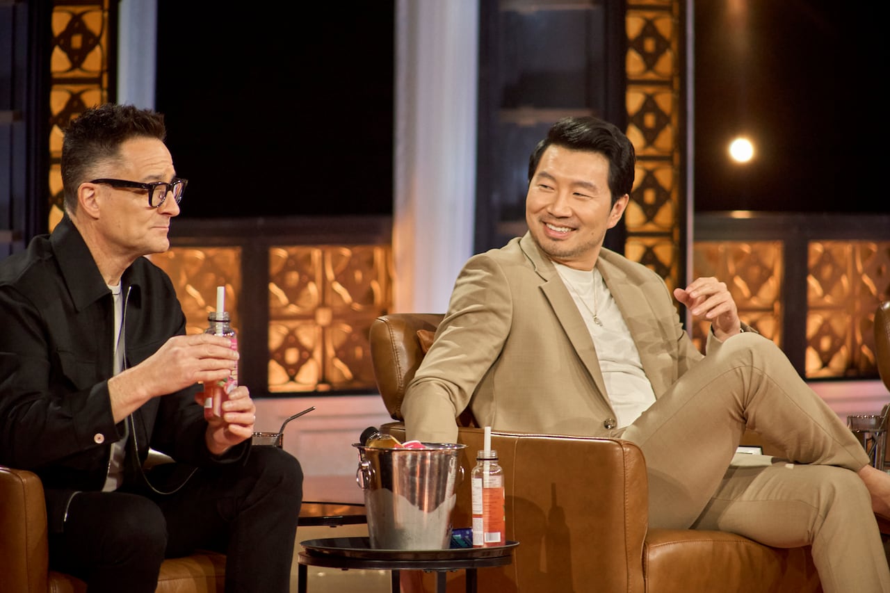 Two men sit next to each other in club chairs, laughing. 