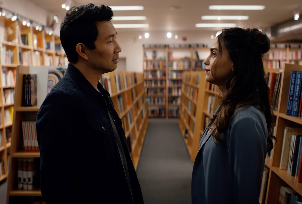 a man and a woman stand facing eachother, staring into eachothers eyes, in a library setting