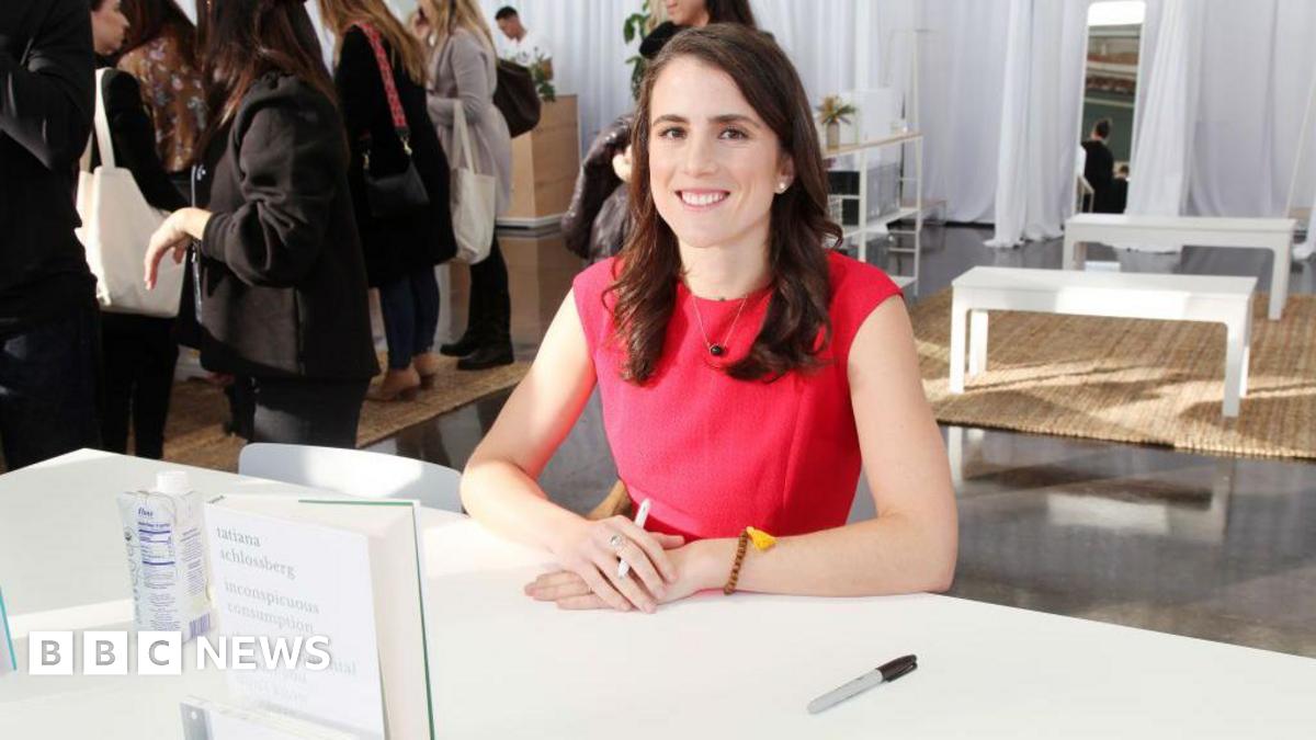 Tatiana Schlossberg with long brown hair and wearing a red short-sleeved dress, sits behind table where her book  is stood up with hands crossed holding a pen