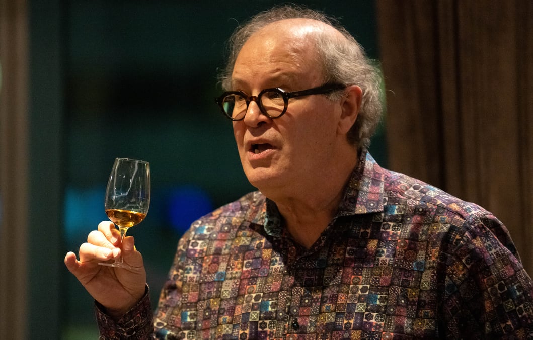 A balding, grey-haired man, wearing glasses and a patterned button-down shirt, speaks as he holds up a small glass containing a sample of brown whisky.