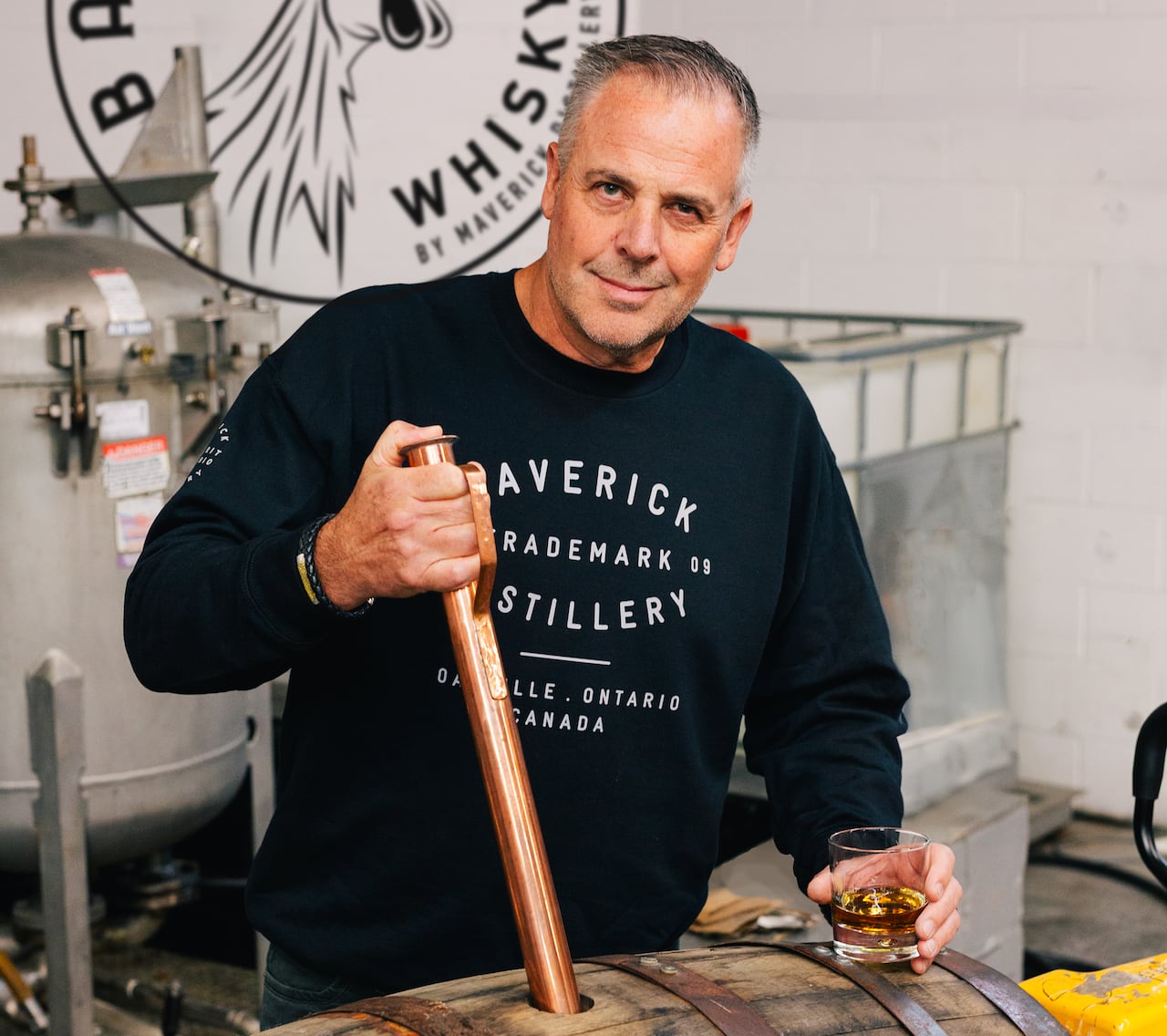 A grey-haired man in a black sweatshirt smiles as he holds a large, circular piece of wood that has been inserted into a hole in the top of a wooden whisky barrel.