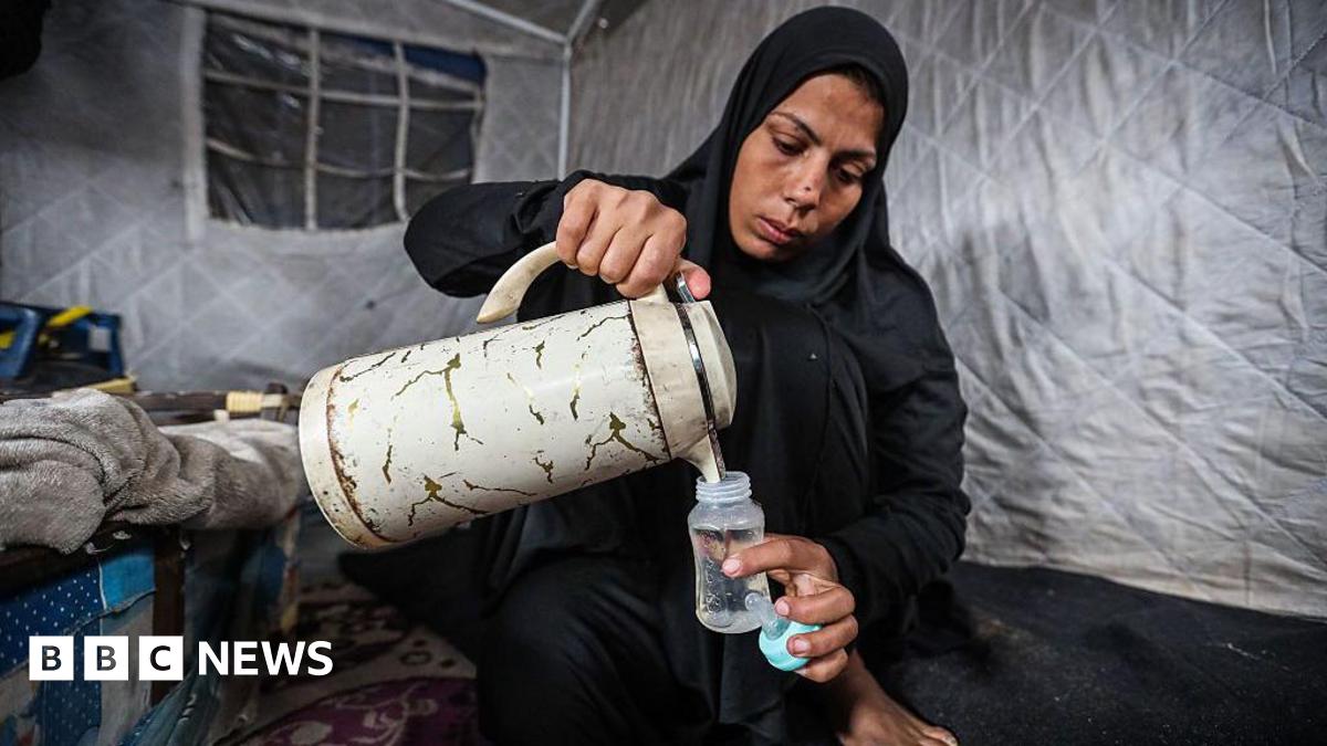 A Palestinian pours water from a large jug into a baby's bottle as she prepares baby formula in a tent